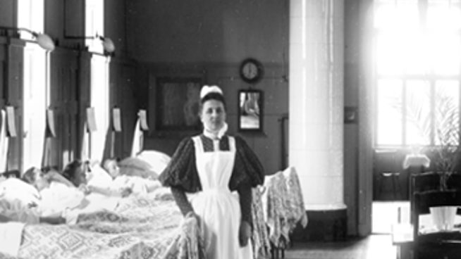 Historiskt svartvitt foto av en vårdklädd kvinna i en stor patientsal med många sängar.