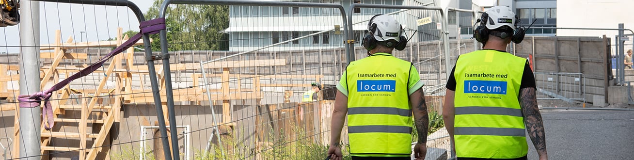 Två män med varselväst och hjälm, bakifrån, på byggarbetsplats.